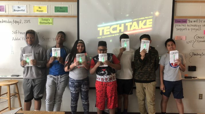 Seven students holding finished paper circuit lightsabers at the front of a classroom.